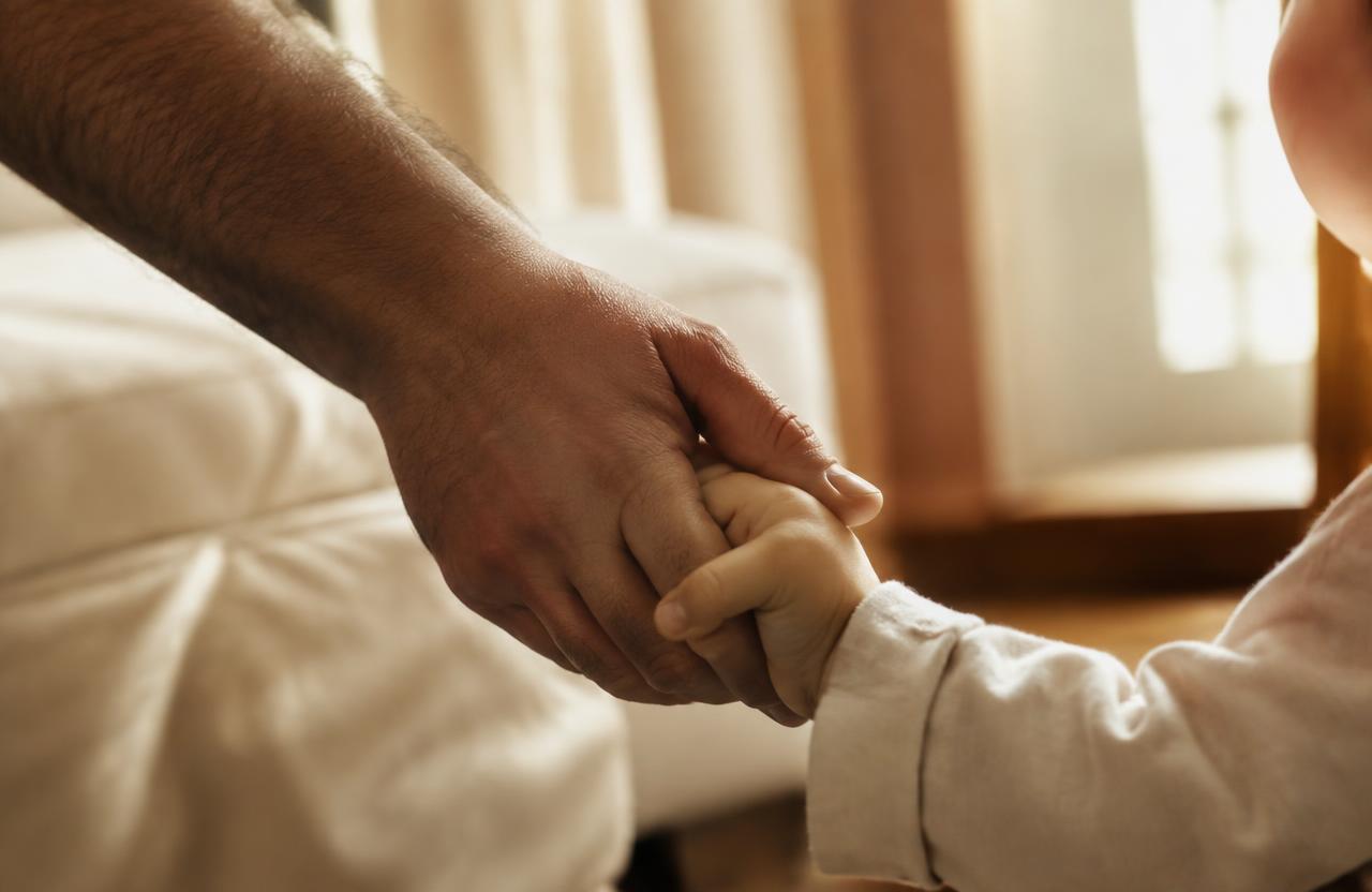 A father holding his small child's hand in soft window light